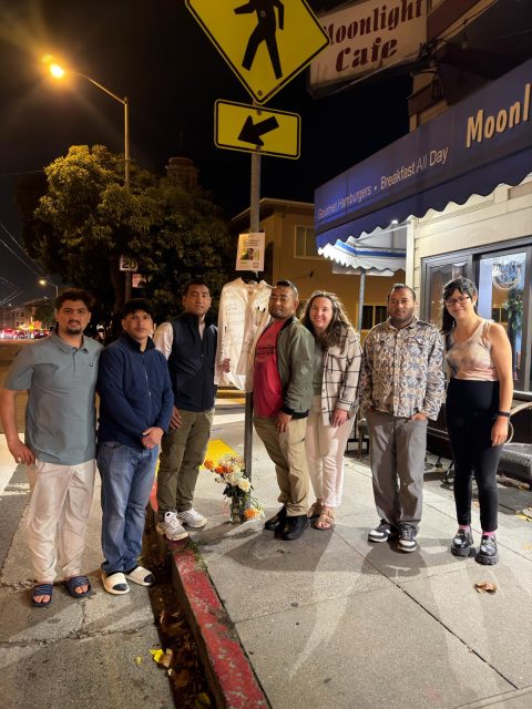 Seven people stand together on a city sidewalk at night under a crosswalk sign, next to flowers and a white shirt memorial outside the Moonlight Cafe.