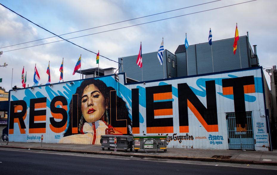A mural on a building features the word "RESILIENT" in large letters, with a painted portrait of a woman. Multiple international flags are displayed on the roof above.