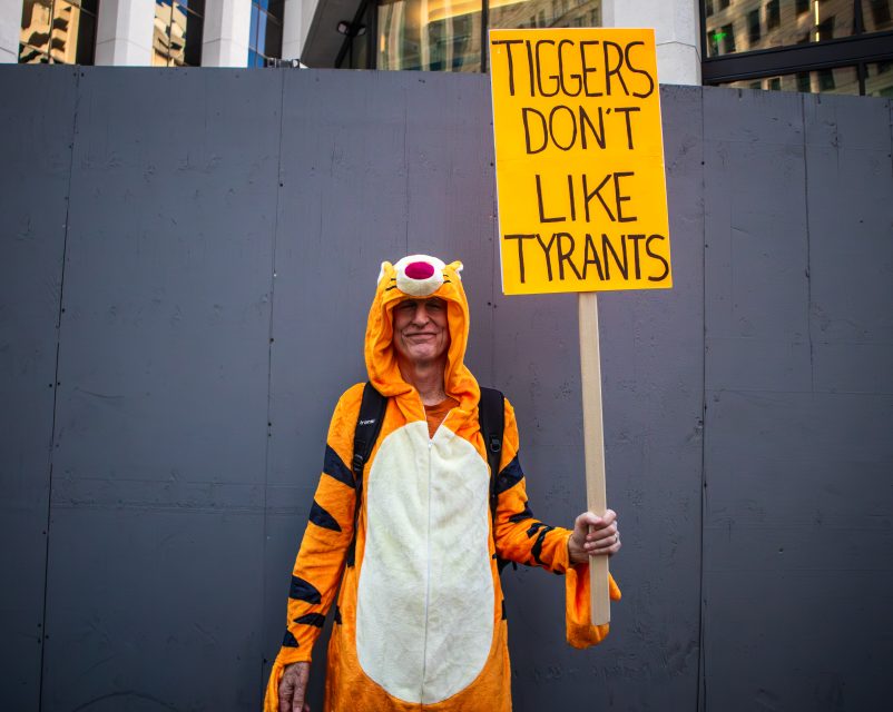 Person in a Tigger costume stands in front of a gray wall holding a sign that reads, "Tiggers don't like tyrants.