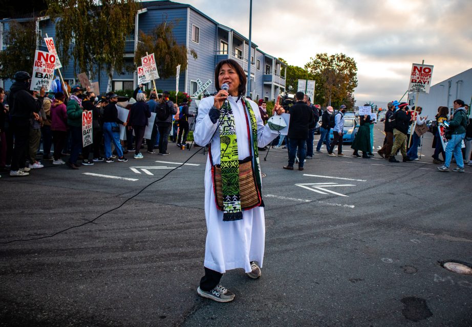 A person in a white robe speaks into a microphone about immigration in front of a crowd holding protest signs on a city street.