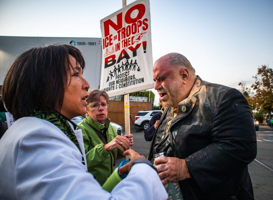 Three people converse outdoors; one holds a protest sign reading "NO ICE TROOPS IN THE BAY" and "Protect Our Neighbors, Defend the Constitution.