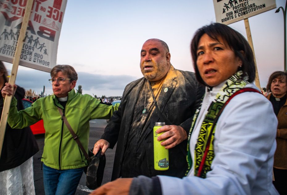 Three people at a protest, one man in the center covered in a yellow powder, are walking together while holding signs and a green water bottle.
