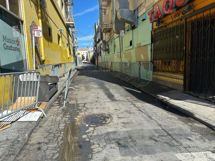 A narrow city alley with metal barricades, cracked pavement, puddles, and buildings on both sides, including a closed storefront and a "Mission Graduates" sign.