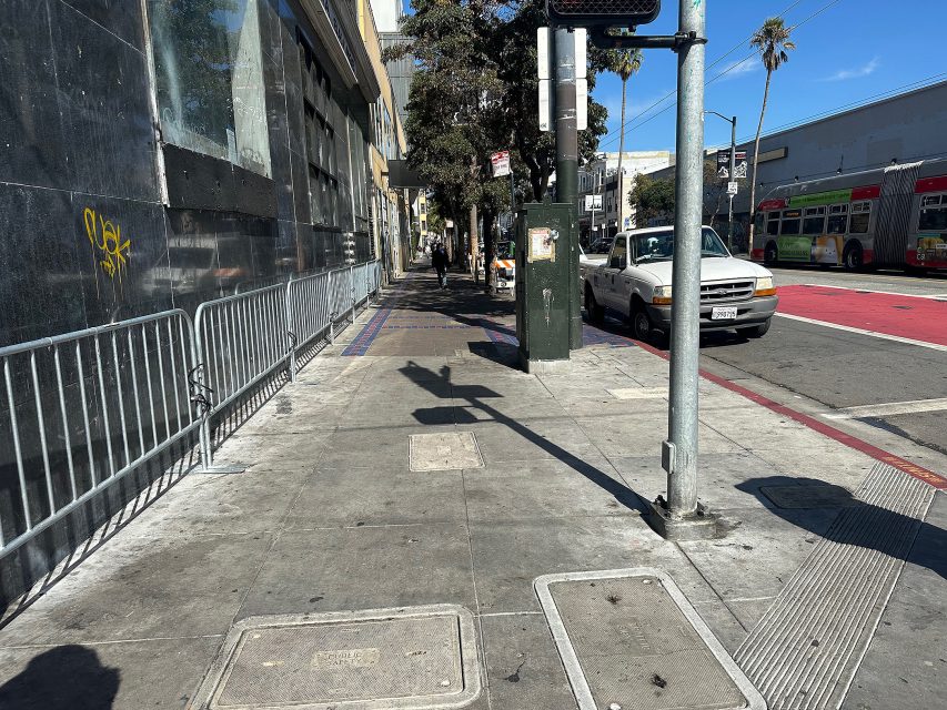 City sidewalk with metal barricades, graffiti on a black wall, a pickup truck, traffic lights, and buses in the background on a sunny day.