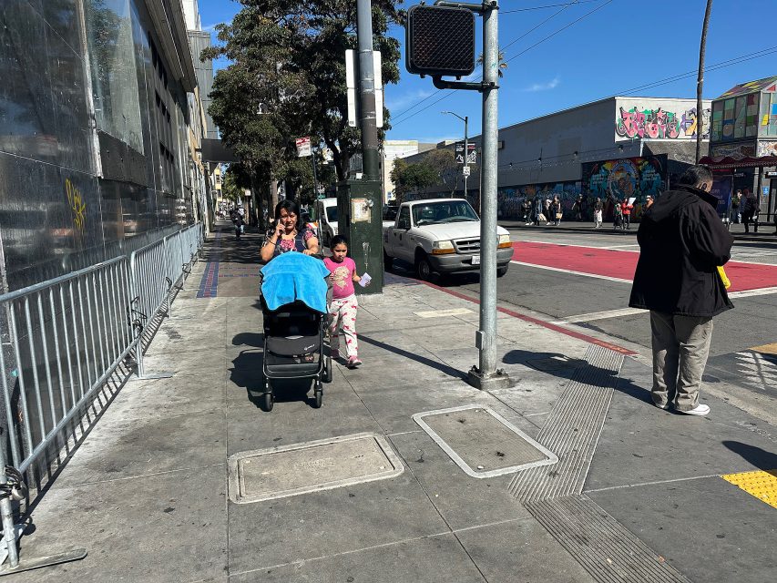 A woman pushes a stroller with two children walking beside her on a city sidewalk. Another person stands near the curb. Cars and street art are visible in the background.