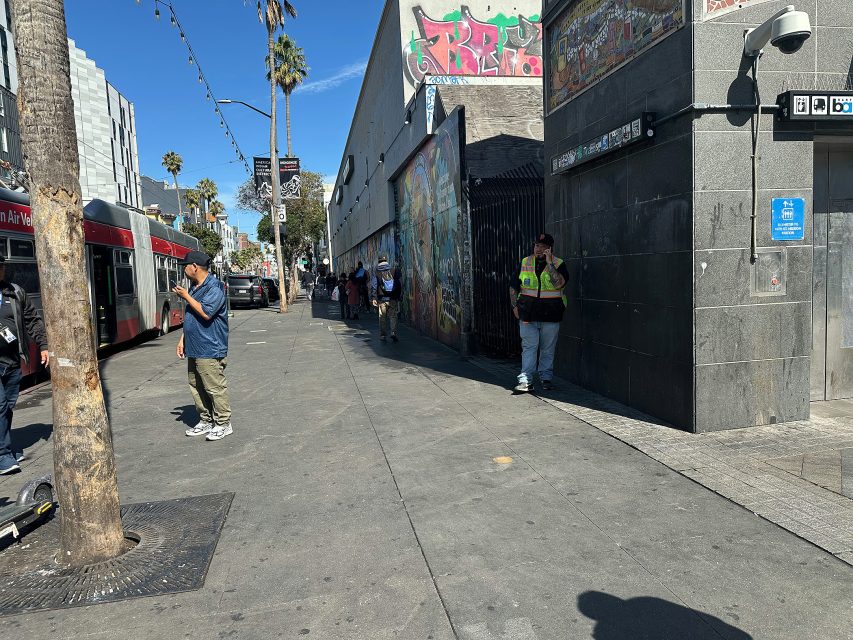 City street scene with several people standing on the sidewalk, a bus on the left, palm trees, graffiti on a building, and bright sunlight creating shadows.