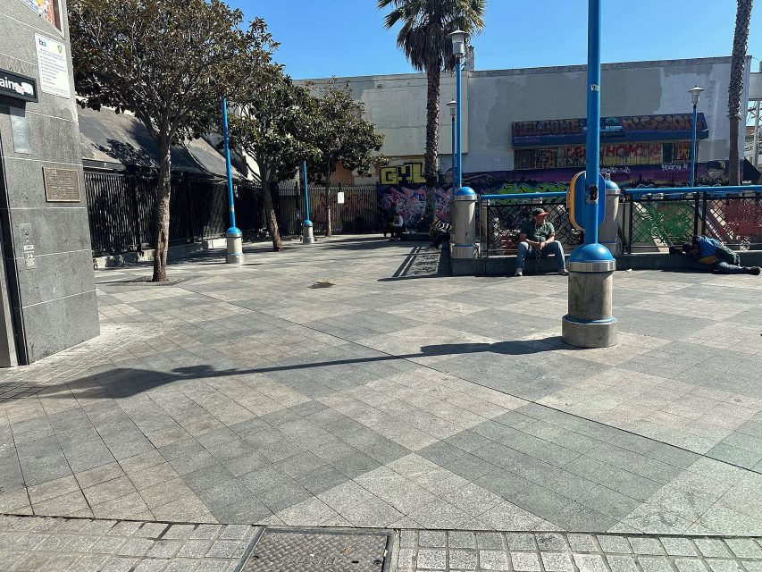 A mostly empty urban plaza with a few people sitting on benches, blue lamp posts, trees, and a graffiti-covered wall in the background.
