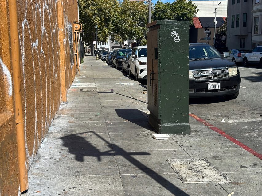 A city sidewalk with cars parked along the curb, a graffiti-covered utility box, and shadows cast by a traffic signal and streetlight.