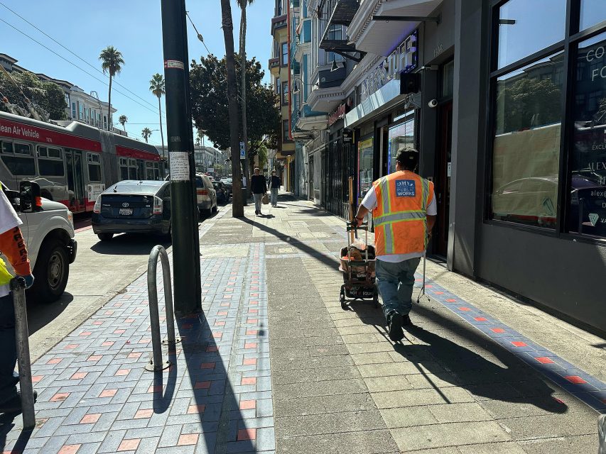 A worker wearing a safety vest pushes equipment along a sunny city sidewalk; a red bus and pedestrians are visible in the background.