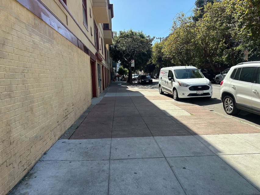 A sunny sidewalk runs alongside a building with parked cars on the street; trees provide partial shade in the background.