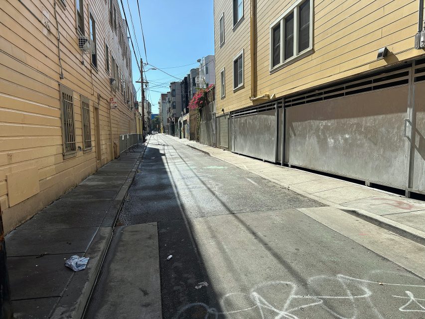 A narrow urban alley with yellow buildings on both sides, graffiti on the pavement, overhead wires, and strong sunlight casting shadows.