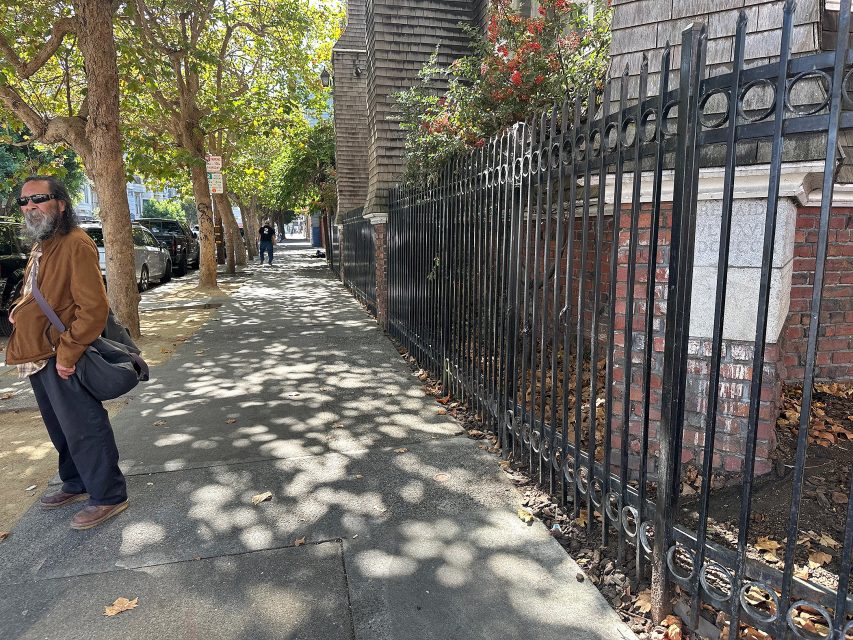 A man wearing sunglasses and a brown jacket stands on a sunlit sidewalk next to a black iron fence and brick building, with trees casting shadows on the ground.