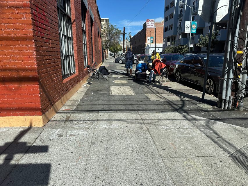 A sidewalk with scattered belongings and a few people near a red brick building; parked cars line the street and a bike is on the ground.