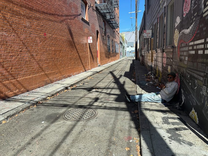 A man sits on the ground against a wall in a narrow, sunlit alley with graffiti, scattered trash, and shadowed brick buildings.