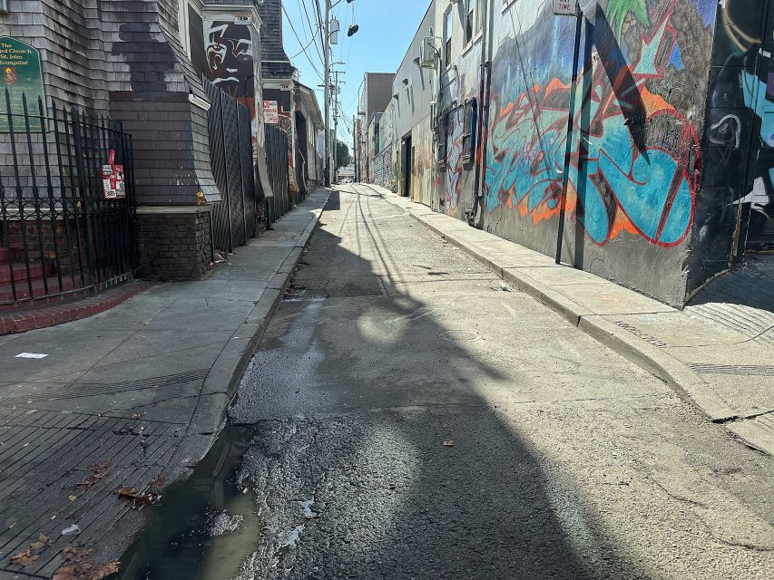 An empty urban alleyway with cracked pavement, puddles, graffiti-covered walls, and overhead utility wires on a sunny day.