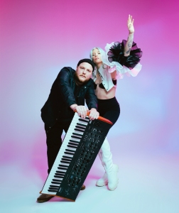 Two people pose in a studio with pink and purple lighting; one leans on a keyboard synthesizer while the other stands with an arm raised, wearing bold, stylish clothing.