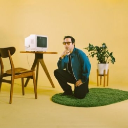 A man in glasses kneels on a green rug in a yellow room next to a table with a vintage computer, a wooden chair, and a potted plant.