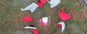 Aerial view of costumes and a scythe arranged in a rough circle on grass. The costumes are mainly red and gray, with one white piece in the center.