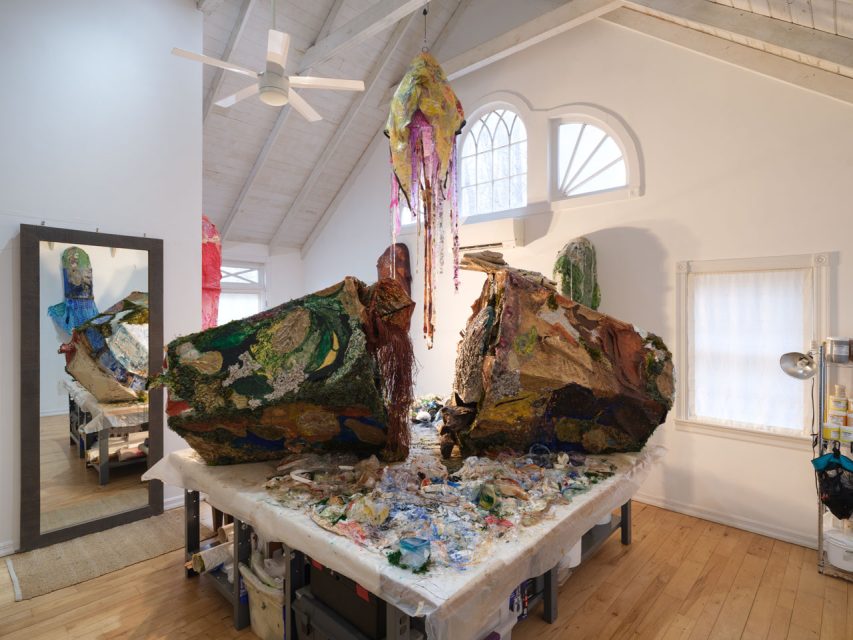Large abstract sculptures made of mixed materials sit on a table in a well-lit art studio with wooden floors, a mirror, and arched windows.