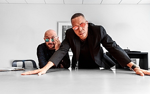Two men with glasses lean over a table in a modern office, one seated and whispering, the other standing with hands on the table, both wearing dark clothing.