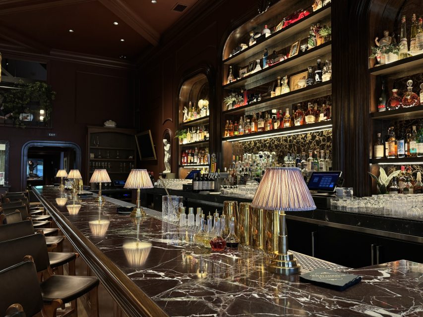 A dimly lit upscale bar with a marble counter, gold lamps, empty glasses, and shelves filled with various bottles of liquor.