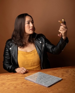 A woman in a yellow top and black leather jacket sits at a wooden table, holding up a small action figure. A notebook with handwritten notes is on the table.