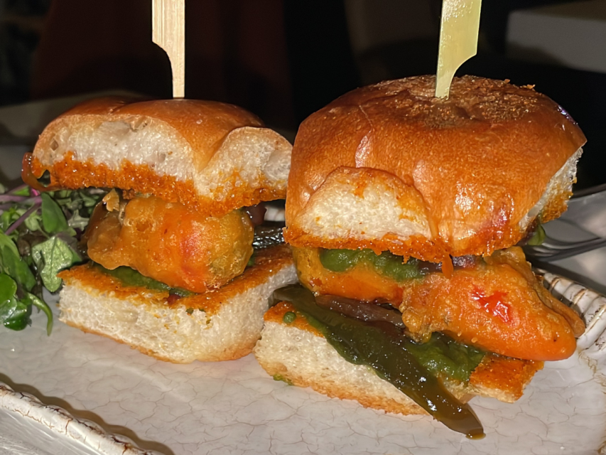 Two vegetable sliders on a plate, each with fried vegetables and greens between toasted buns, secured with wooden picks.