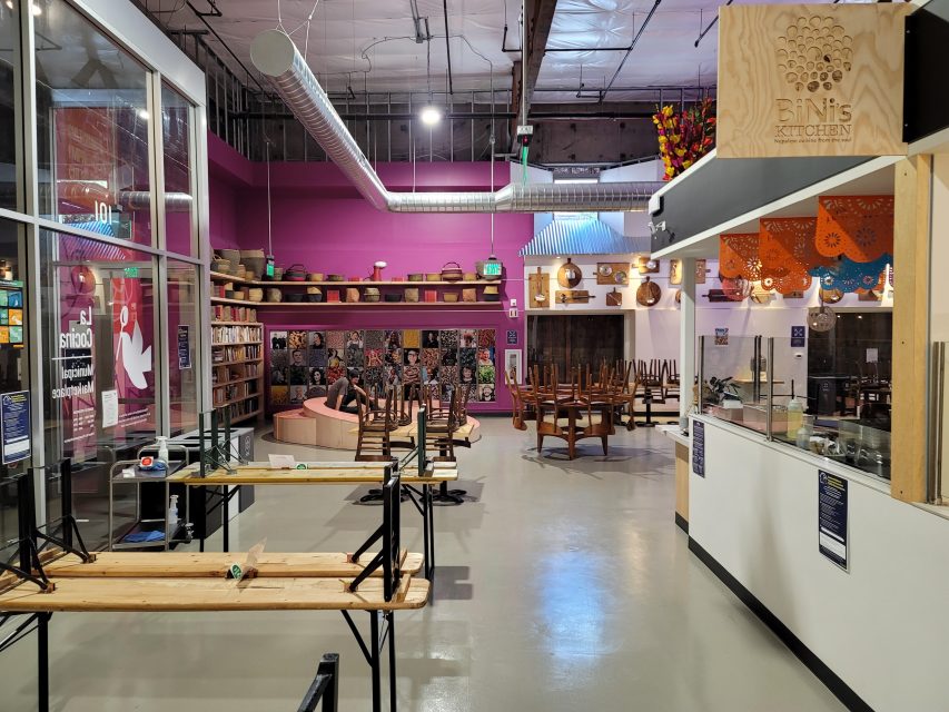 Interior of a food hall with stacked chairs on tables, a seating area with shelves and baskets, and a purple accent wall in the background.