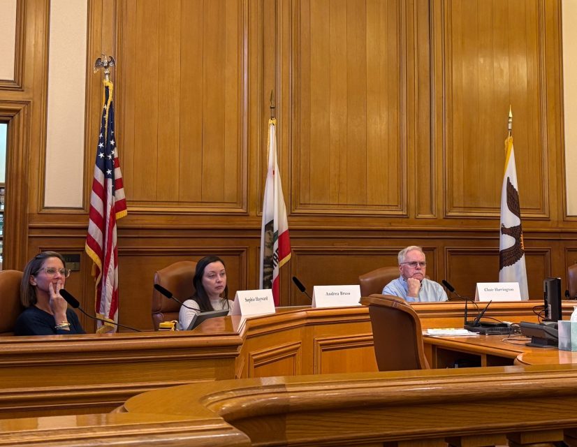 Three people sit at a wooden panel desk in a formal meeting room with microphones, nameplates, and flags in the background.