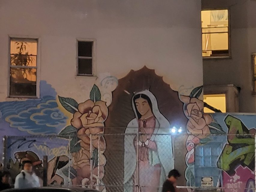 A mural of the Virgin of Guadalupe surrounded by roses is painted on a building wall, partially obscured by a chain-link fence, with people walking by at night.