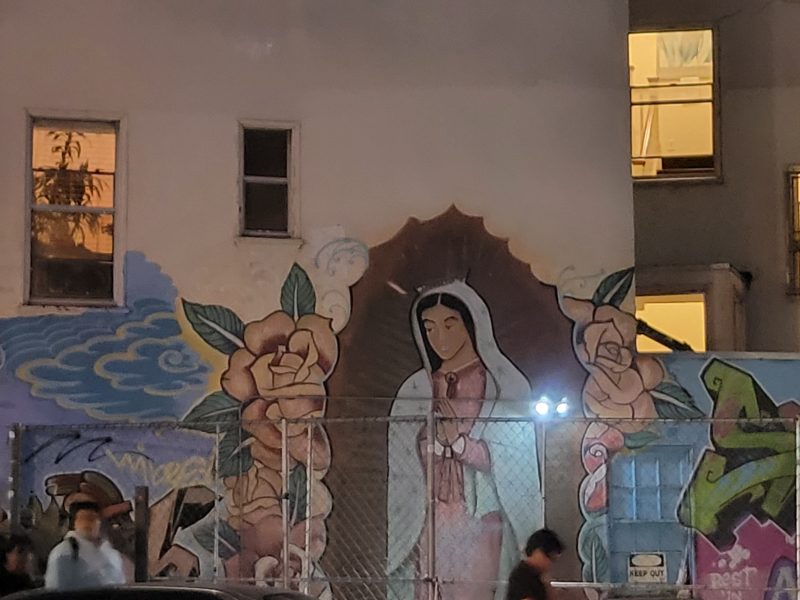 Mural of the Virgin Mary with roses and clouds painted on a building wall behind a chain-link fence at night; people walk by in the foreground.