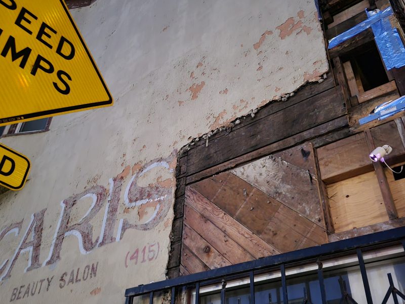 A faded beauty salon sign and weathered wooden boards on a worn building exterior, with a yellow "Speed Humps Ahead" street sign overhead.