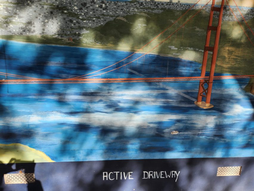 A mural of the Golden Gate Bridge over blue water, with the words "ACTIVE DRIVEWAY" painted at the bottom and tree shadows falling across the image.