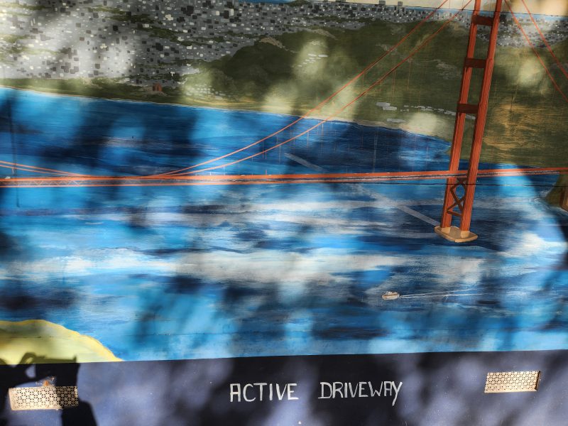 Mural of the Golden Gate Bridge over water with the words "ACTIVE DRIVEWAY" painted at the bottom; shadows of leaves are visible on the mural.