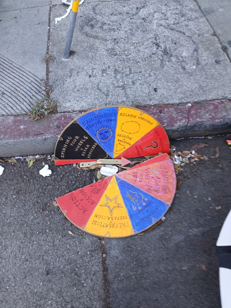 A broken, colorful cardboard spinning wheel with various actions and phrases lies on a dirty sidewalk near a curb.