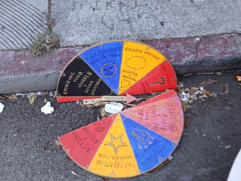 A broken, colorful cardboard spinning wheel with various actions and phrases lies on a dirty sidewalk near a curb.