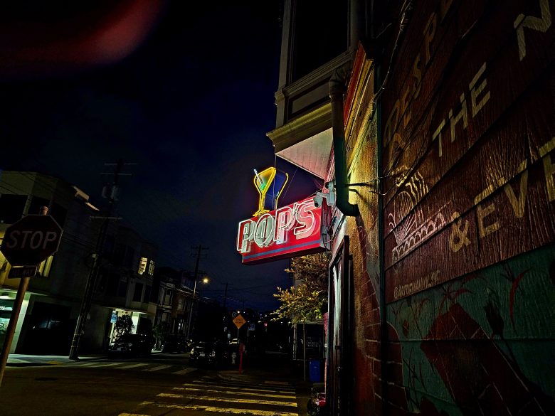 Neon sign reading "POPS" with a martini glass graphic glows above a sidewalk on a dark, empty city street at night.