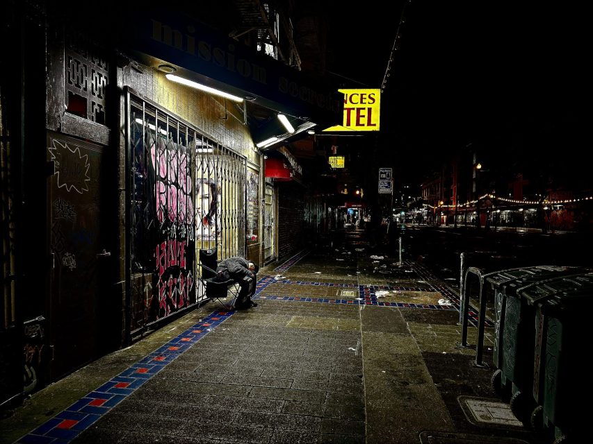 A person crouches on a wet sidewalk at night near a gated storefront with graffiti, under bright lights on a mostly dark, empty street.