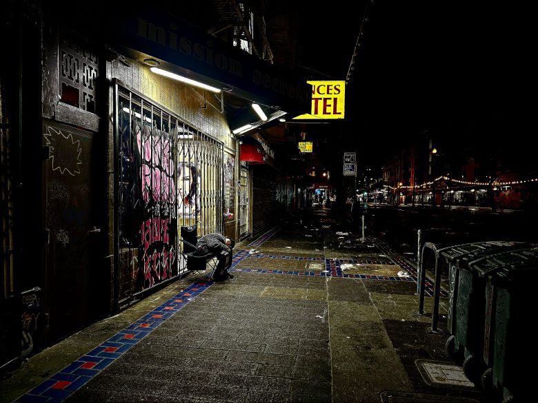 A person crouches near a gated storefront on a dimly lit urban street at night, with neon signs and wet pavement visible.