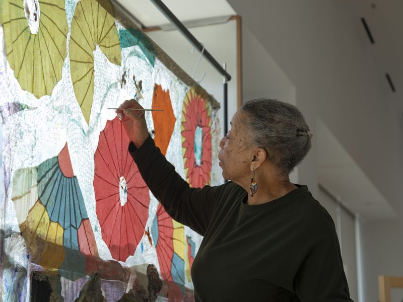 An older woman stands in front of a colorful artwork with umbrella motifs, painting details onto its surface with a small brush in a well-lit indoor space.