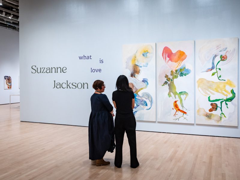 Two people stand in an art gallery viewing a colorful triptych painting by Suzanne Jackson, with the text "Suzanne Jackson what is love" on the wall beside them.