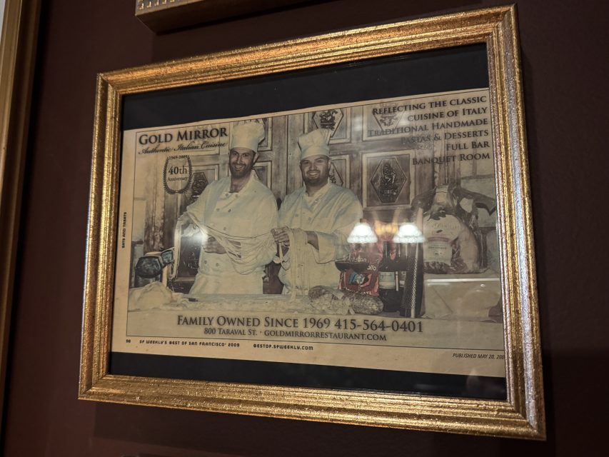 Framed photograph shows two chefs in white uniforms and hats standing in a restaurant kitchen, with a printed advertisement for Gold Mirror Restaurant below them.