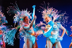 Two performers in elaborate feathered costumes and headdresses dance and smile on stage under bright lighting.