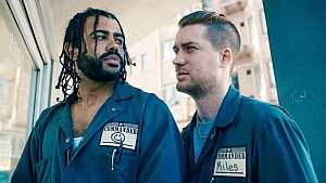 Two men wearing matching work uniforms with name tags stand side by side, looking in the same direction outside a building.