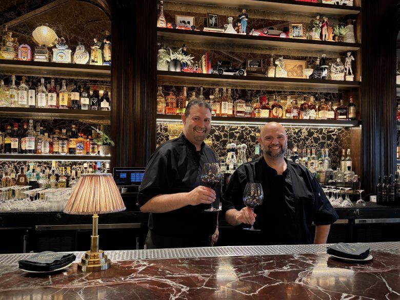 Two chefs stand behind a marble bar, holding wine glasses and smiling. Shelves behind them display various bottles and decorative items. A small lamp sits on the bar.