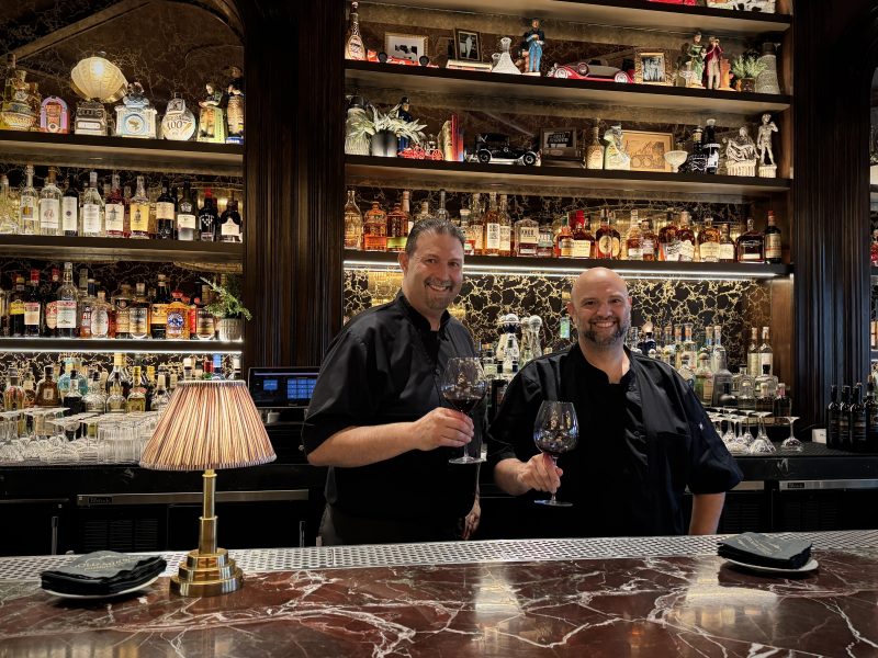 Two chefs stand behind a marble bar, holding wine glasses and smiling. Shelves behind them display various bottles and decorative items. A small lamp sits on the bar.