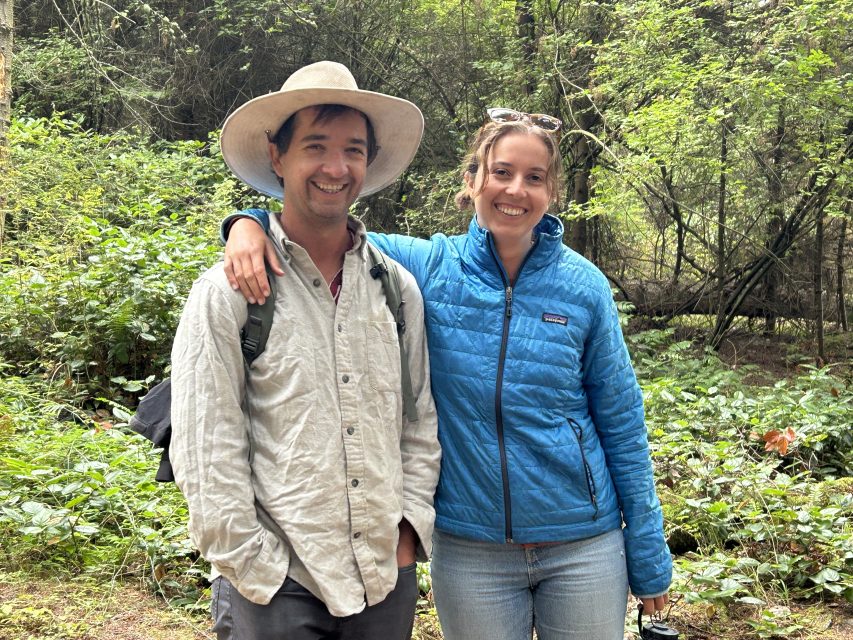 Two people standing side by side in a forest, one wearing a wide-brim hat and the other in a blue jacket, both smiling at the camera.