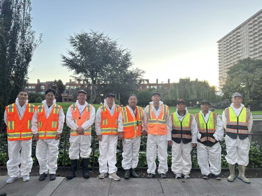 Ten workers wearing white coveralls and high-visibility vests stand in a row outdoors, with trees and buildings in the background.