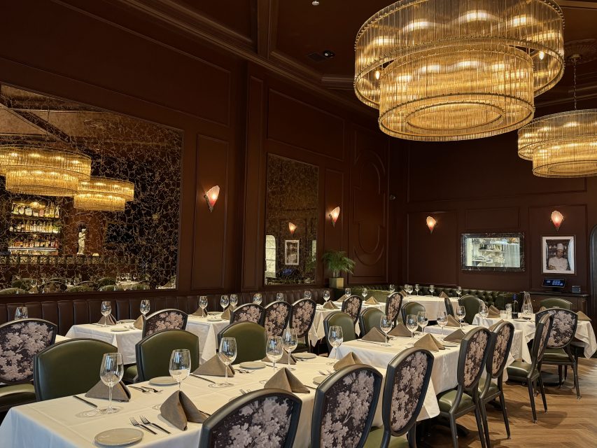 Elegant restaurant dining room with white tablecloth-covered tables, set with glassware and napkins, ornate chandeliers, large mirrors, and brown paneled walls.