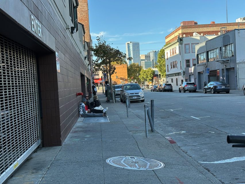 Several people sit on the sidewalk beside a building in an urban area, with cars parked along the street and city buildings visible in the background.